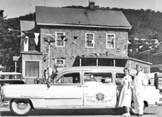 Black and white photograph of a man and woman standing beside a 1950s ambulance with buildings and mountains in the background